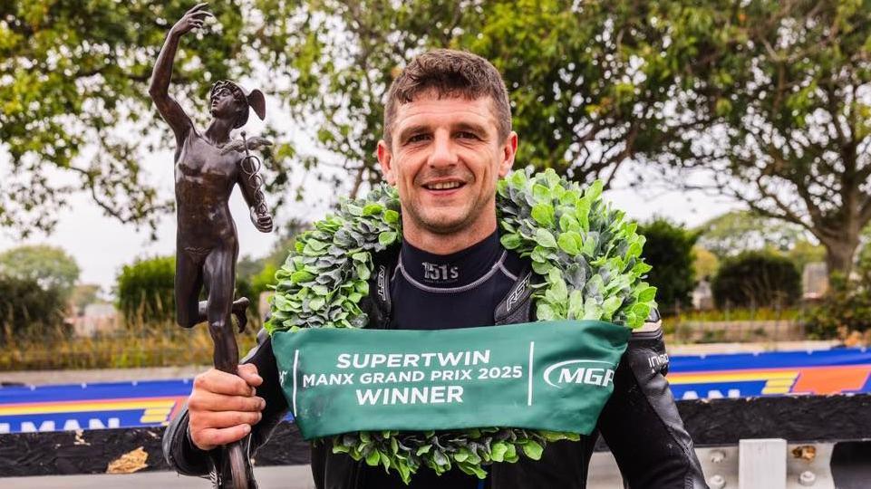 Lewis Arrowsmith, who is wearing black leathers and has a laurel garland around his neck with a green banner that says Supertwin Manx Grand Prix Winner across it. He is holding a bronze MGP trophy.