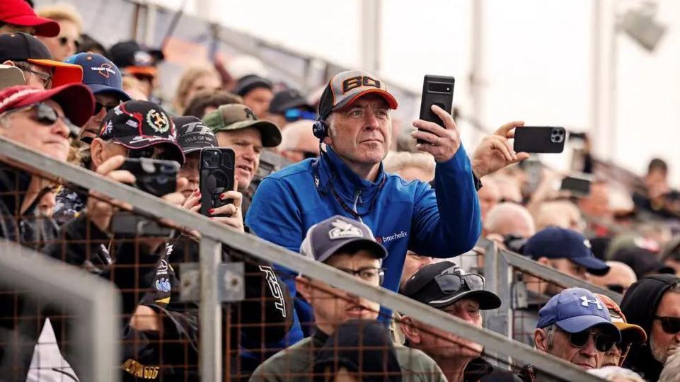 Race fans sitting on the grandstand holding up mobile phones to film the bikes.