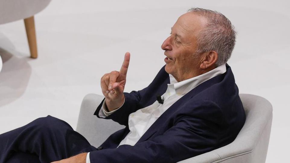 Larry Summers, sat in profile on a grey armchair, wearing a navy blue suit and white short, smiles as he raises the index finger of his right hand