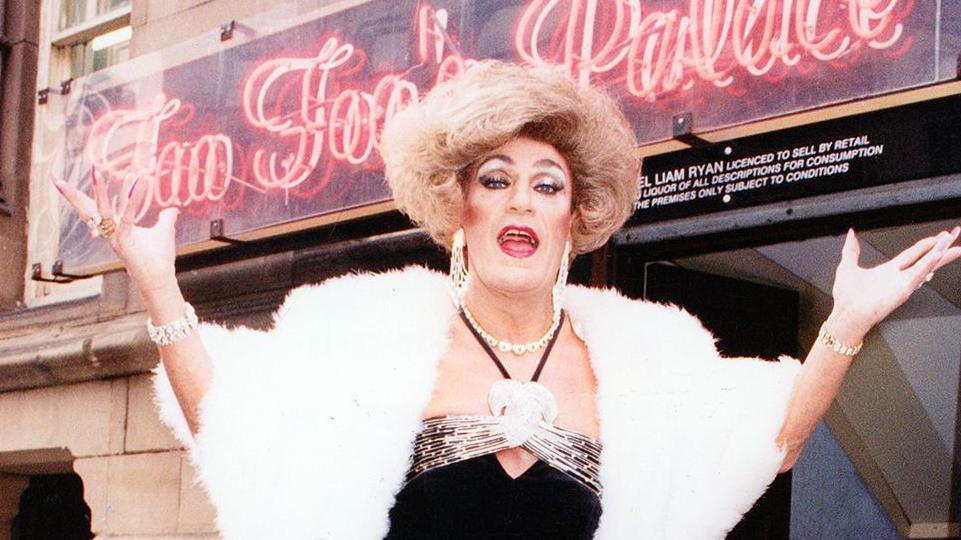 Frank Pearson, performing as drag queen Foo Foo Lammar in a blonde wig, make-up and a black evening dress. Photographed outside the entrance of Foo Foo's Palace nightclub in Manchester, the entertainer wears dangly earrings, a pearl necklace and a white faux fur bolero jacket.