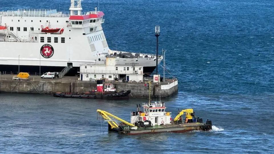 A small dredging boat is in Douglas Harbour. It has a small yellow crane-like device at one end and another piece of yellow equipment at the other. The Manxman ferry is visible on the other side of the pier.