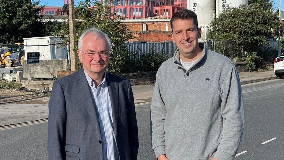 Jeremy Hilton and Luke Shervey in front of the site of the former Wessex House in Gloucester. Mr Hilton is wearing a grey blazer and blue striped shirt. He has short grey hair and glasses. Mr Shervey is standing next to him. He has short dark hair and is wearing a grey jumper and black jeans. Both men are looking at the camera and smiling. Behind them, cranes and building equipment and vehicles can be seen. 