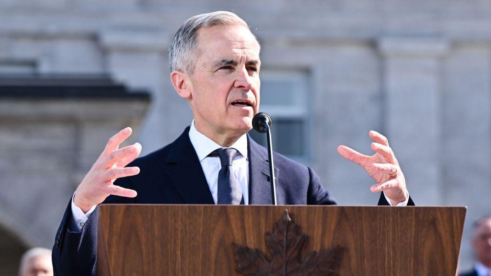 Mark Carney is a white man with grey hair. He gestures with his hands as he speaks behind a wood podium with a Canadian Maple Leaf symbol on the front