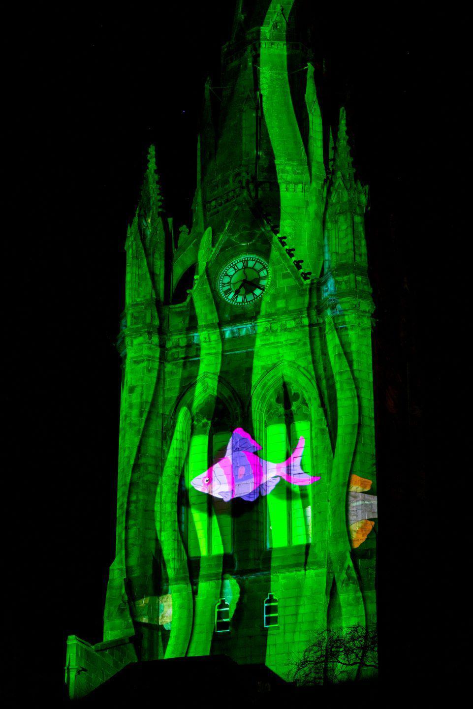 Clock tower lit up green, with a pink fish 'swimming' across it.