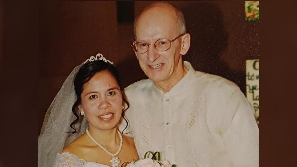 Paul and Juliet Adams, pictured on their wedding day. Juliet (left) is wearing a tiara and veil, pear earrings and necklace and a white lace dress. She has dark hair and is smiling while holding a bouquet. Paul (right) is also smiling, and wears round glasses and a white shirt with a short round collar.