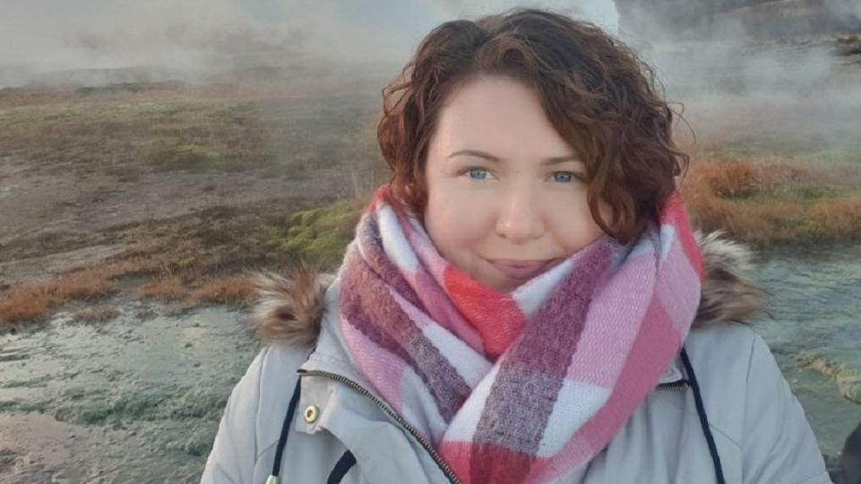 Emma Drinkwater wearing a pink and white checked scarf and a grey coat. She has brown curly hair and is smiling at the camera. She has blue eyes.