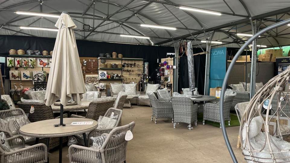 A covered outdoor area of a garden centre, with six brand-new sets of grey wicker patio tables, chairs, and parasols. The walls of the room are black, and hung on the far wall are various pieces of garden wall art including wrought iron sculptures of butterflies and frogs.