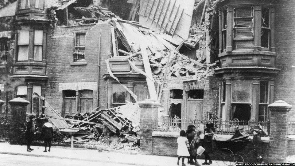 A black and white photograph of two groups of children looking at destroyed houses in a Hartlepool street. One of them is leaning on a pram.