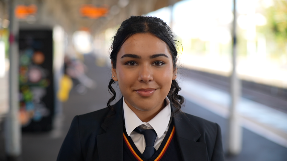 Nishaal a girl with dark curly hair, smiles at the camera. She is wearing a dark-coloured blazer and dark tie as part of her school uniform.