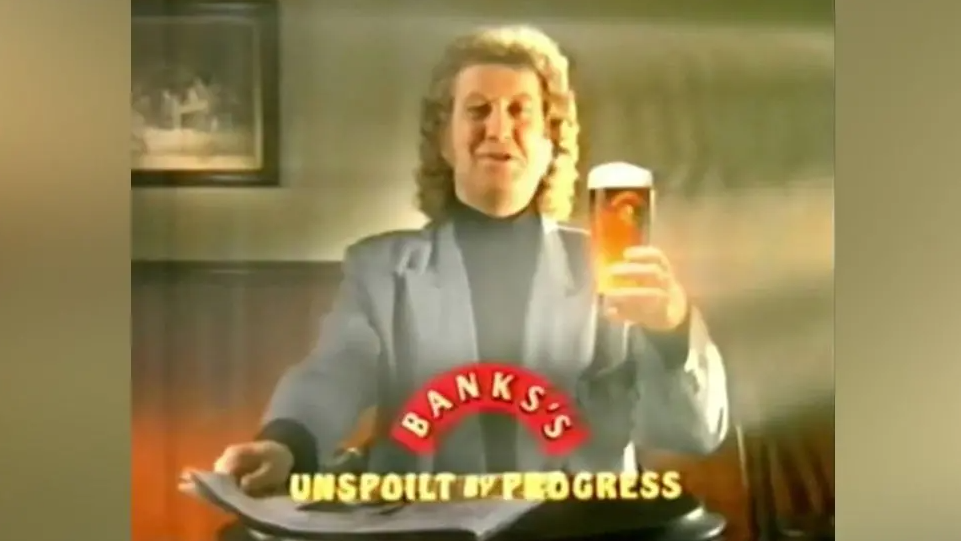 Noddy Holder is sat at a pub table holding a pint with a newspaper in a TV advert for Banks's. He is wearing a grey suit and black jumper.
