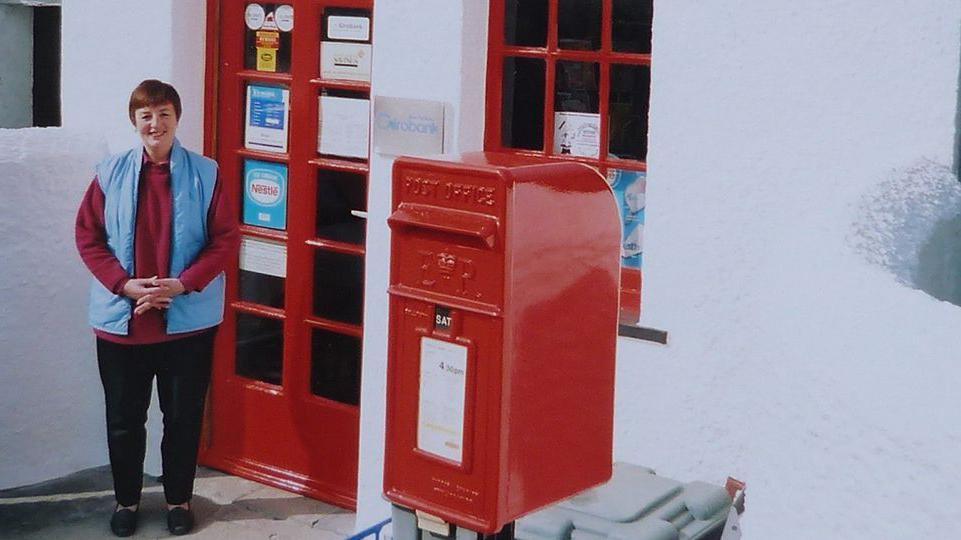 A picture of Alun's wife, Evelyn outside their post office in Llanfarian, near Aberystwyth. The picture looks like it was taken many years ago before the business closed. She is smiling. 