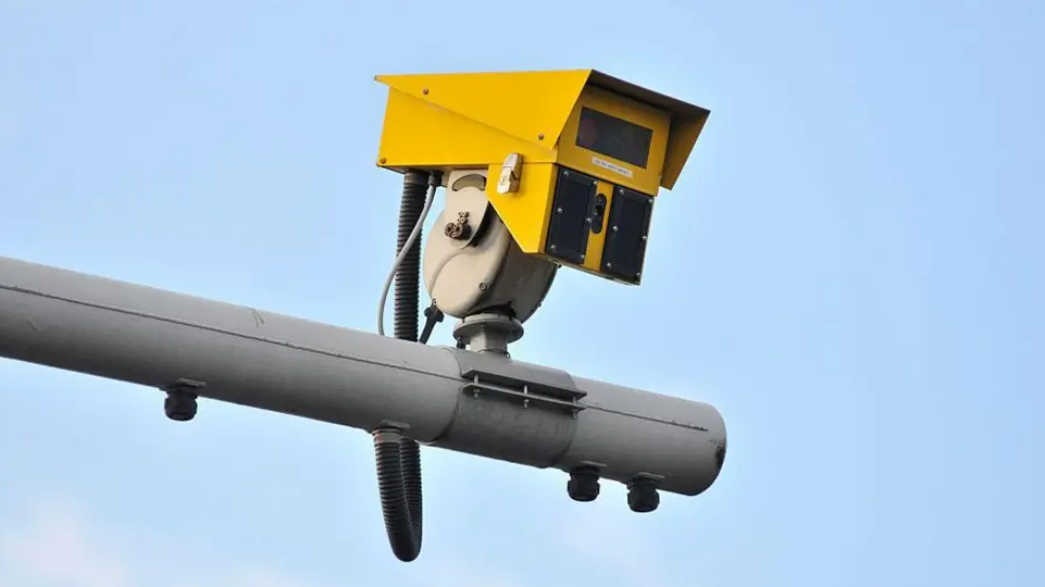 A speed camera can be seen. It is yellow and is attached to a grey pole with a black cable coming out out of it and attaching to the pole.
