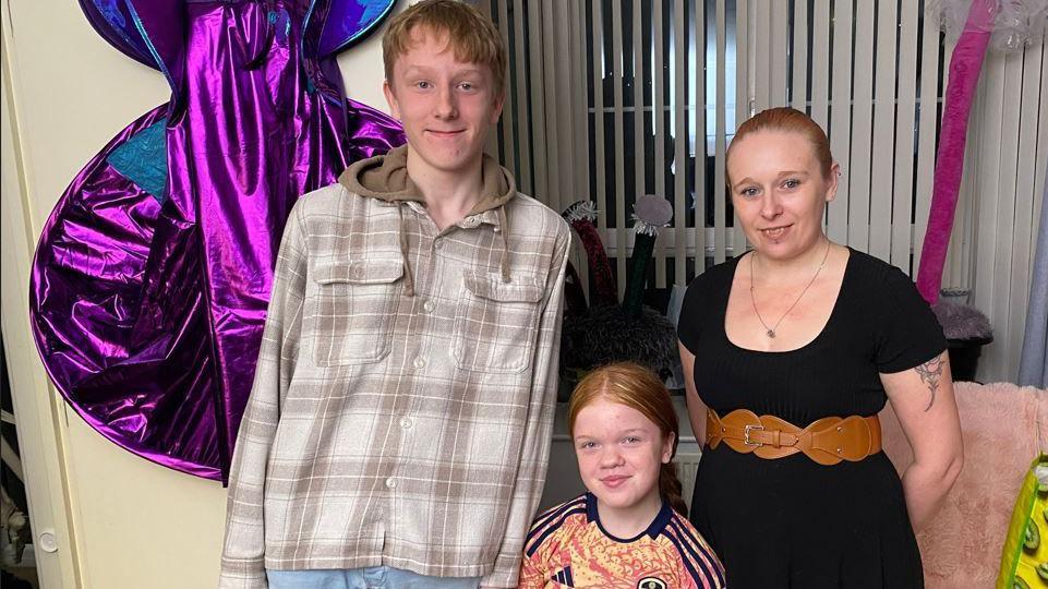 Susan with her children Brandon and Jessica standing in the living room of their home. A purple and blue halloween costume with a cape is hanging on the door