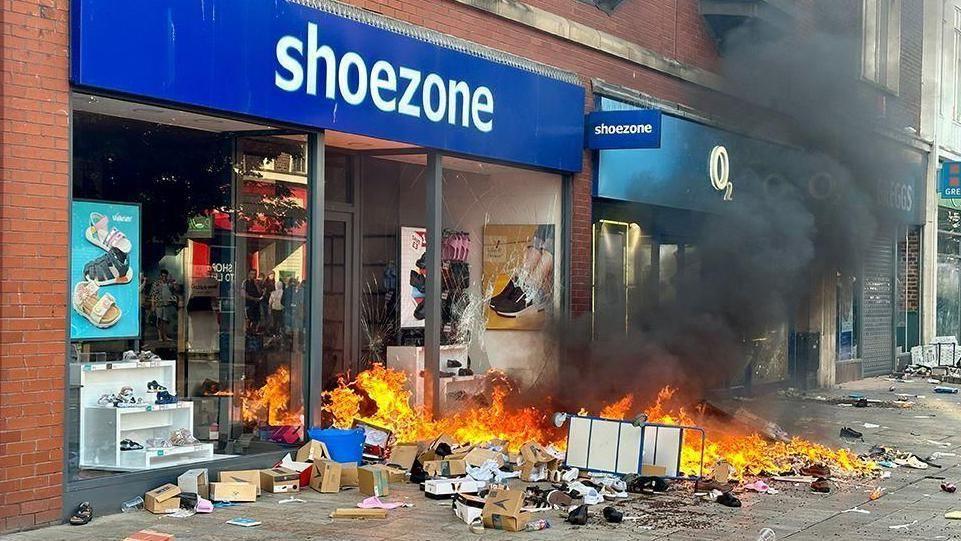 Objects and other items on fire in front of Shoezone shop with black smoke billowing from the strewn rubbish on the ground