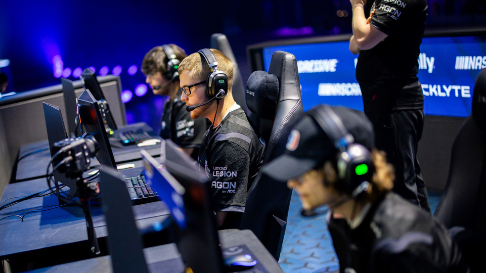 Esports players take part in a competition. They are sat at desks and laptops sitting in black seats wearing black tops with headphones on.