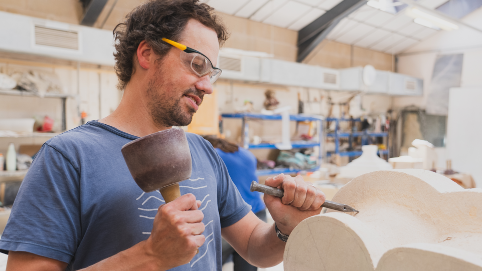 A stonemason wearing safety glasses in a large workshop holding a chisel and hammer and carving a piece of stonework. There are lots of blurred shelves of tools and other equipment in the background.