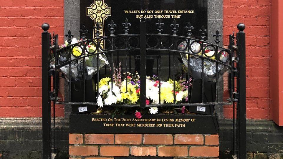 A memorial printed on a black plaque with gold lettering. Flowers are sitting around the memorial. A small black fence surrounds the plaque.