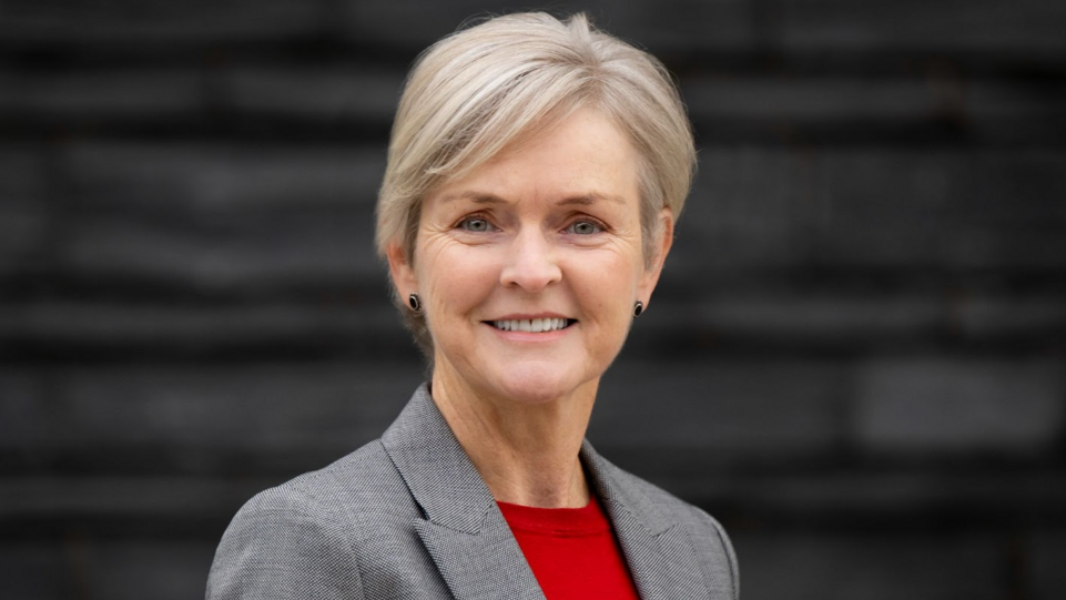 Delyth Evans wearing a grey jacket and red top, in front of a slate background which is out of focus. She has short blonde hair and is wearing a pair of black circle stud earrings while smiling at the camera.