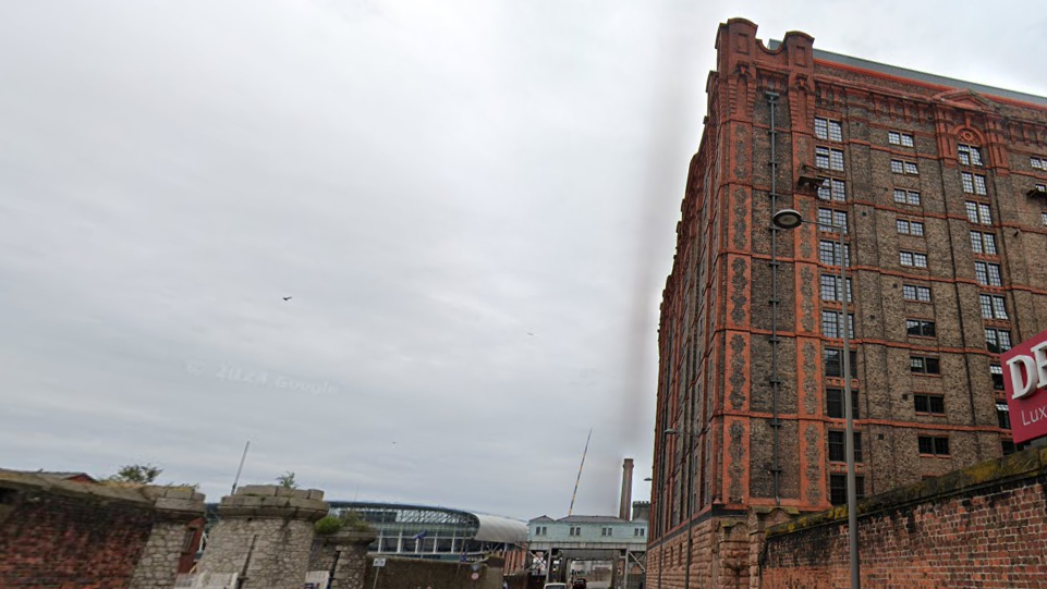 Image shows the redeveloped Tobacco warehouse on the Liverpool docks. It is a vast brick building more than 10 storeys high. To the left-hand side of the image, the Everton Hill Dickinson Stadium can be seen in the distance.  