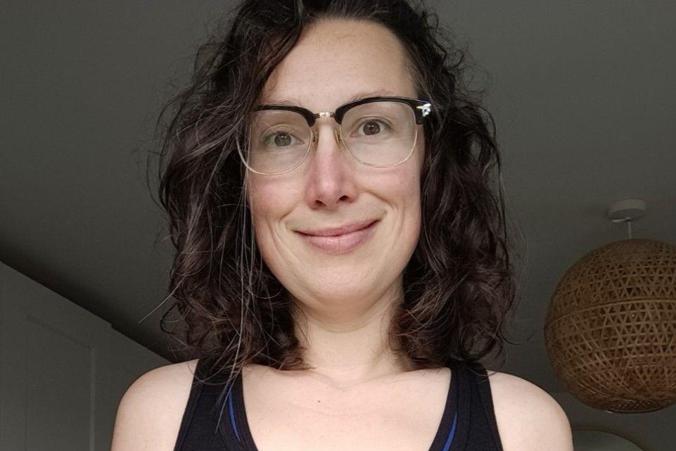 A woman with dark hair, a black top and glasses smiles at the camera. She is stood in a house