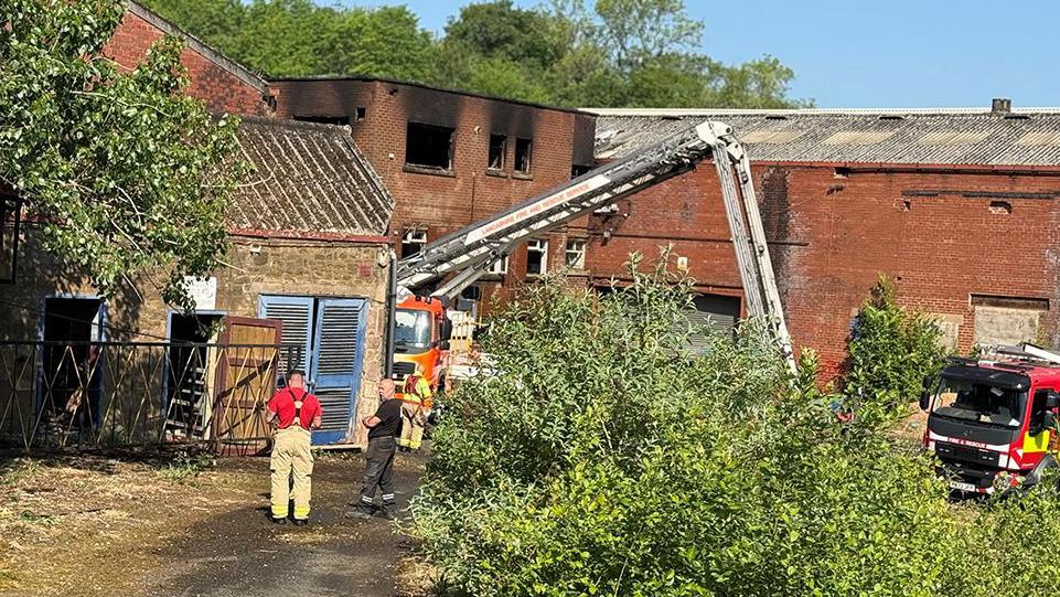 Fire fighters and an aerial ladder by the derelict mill, a red brick building with blackened windows where the fire has been put out