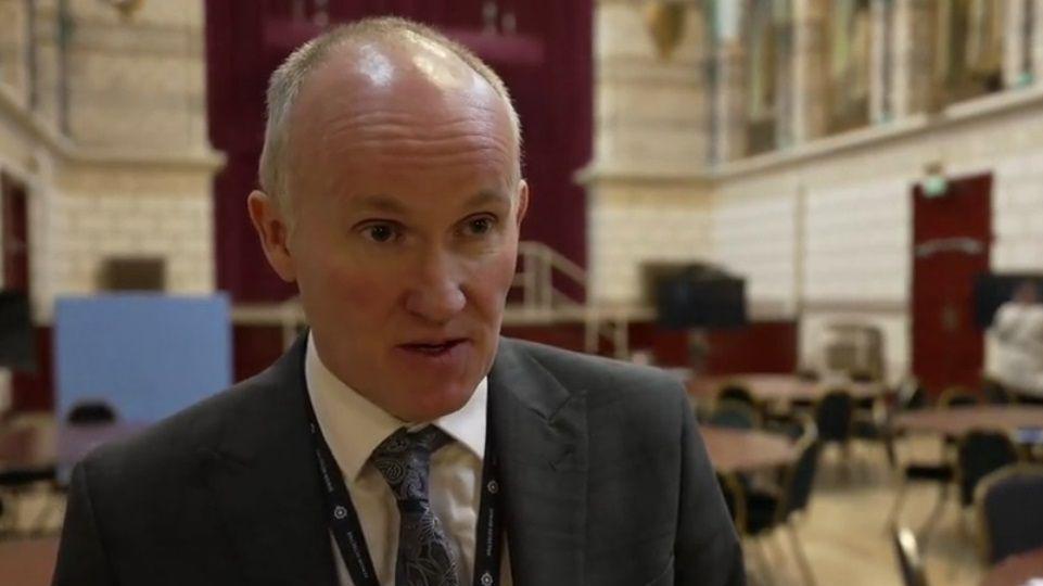 Det Supt Glenn with short fair hair wearing a grey jacket and blue tie with a blue lanyard.  There are tables and chairs behind him as well as a stage.