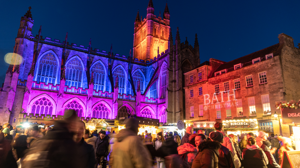 Bath's Georgian architecture is lit up by lights, with market stalls spread out at Christmas. There is a large crowd walking around, under the night sky.