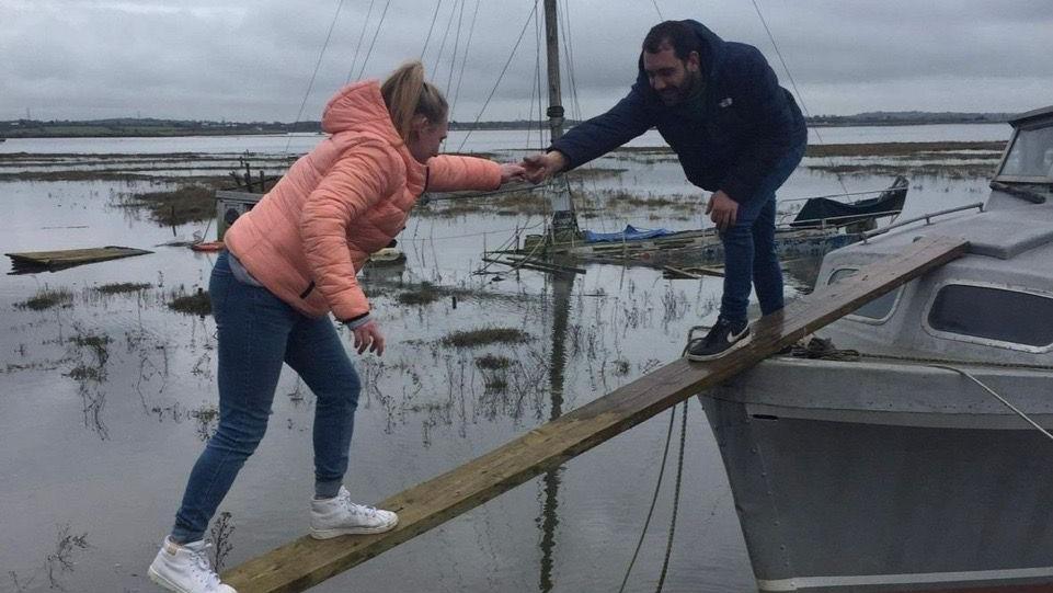 A man and a woman using a wooden plank to get onto a boat. She is wearing jeans and a pink coat. He is wearing jeans and a navy coat.