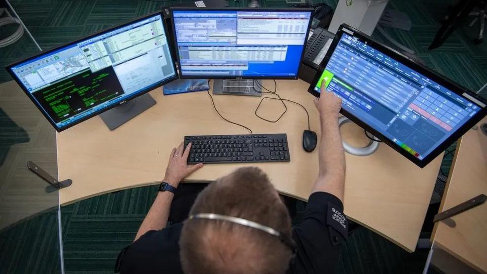 The top of a man's head who's wearing a headset and is looking at three screens with information on them and he has a hand on a keyboard and another on a touch screens