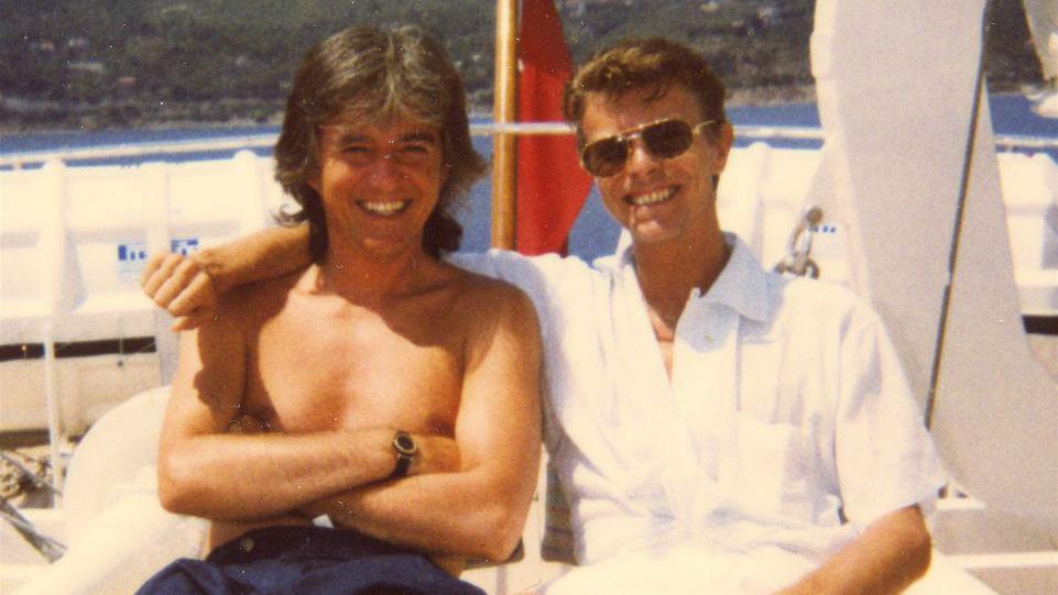 George Underwood and David Bowie sitting on the deck of a boat with the sea and land behind them