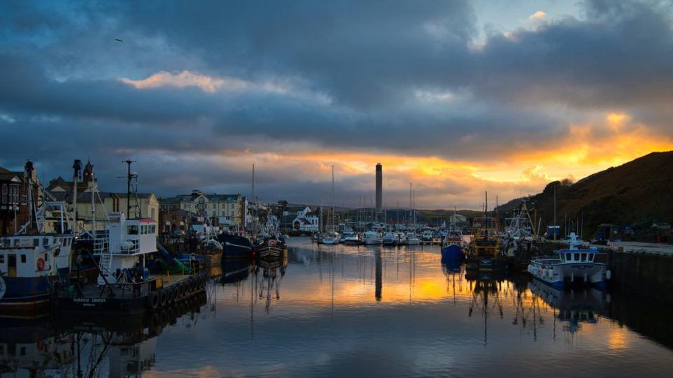 Peel Marina, the water is still with fishing boats and sailing boats moored against the harbourside and pontoons at sunset.