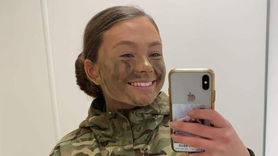 A mirror selfie of Gunner Beck wearing her camouflage military uniform, with her hair tied back into a slick low bun. She has camouflage green and brown face paint on and is smiling at the mirror.