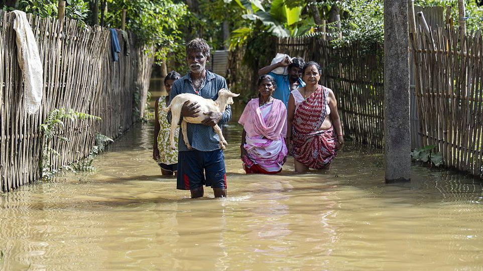 Residents - two men and three women - wade through floodwaters while carrying a goat and salvaged belongings from their inundated homes in a flood-affected neighborhood, on July 22, 2026, in Jorhat, India