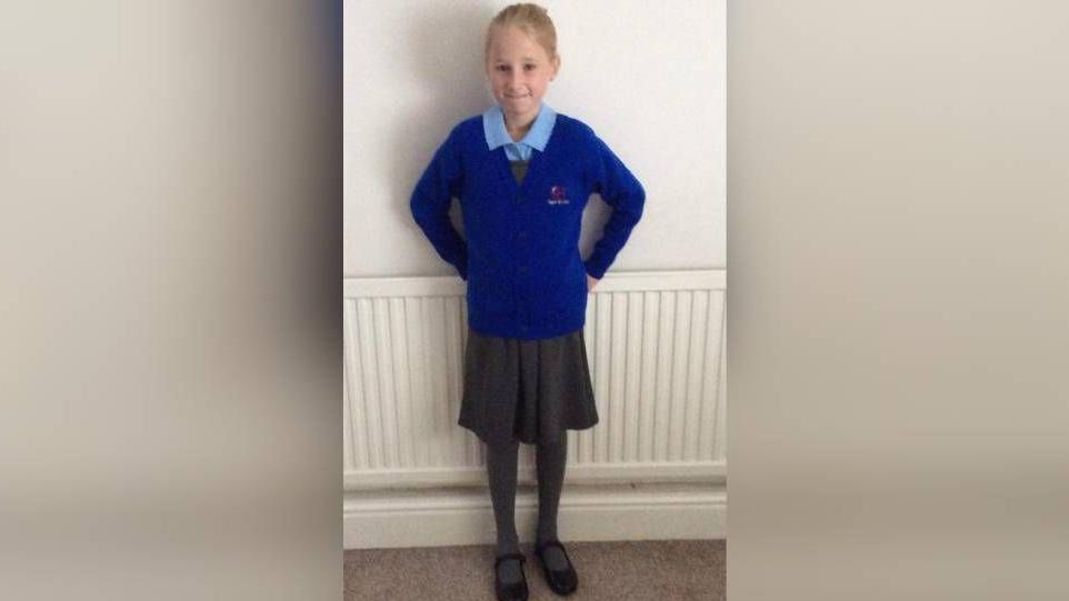 A young girl with blonde hair wearing a blue school uniform, a school skirt and black school shoes. She is stood in front of a radiator leaning on a white wall.