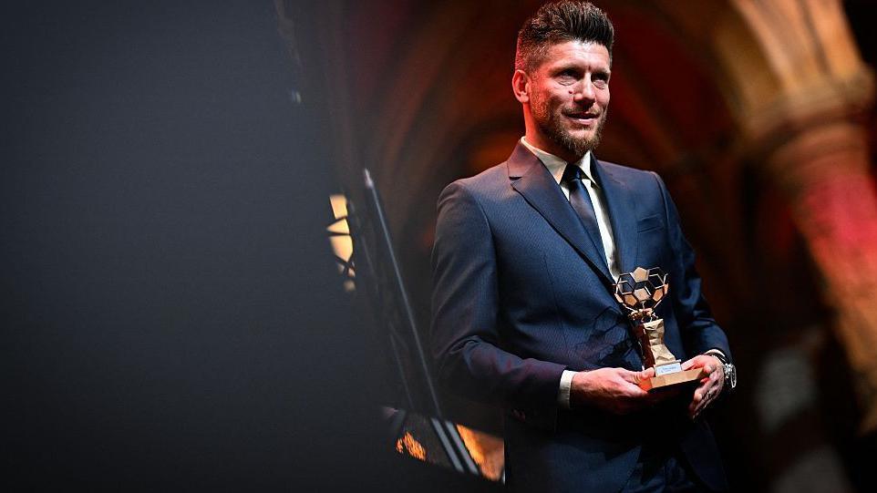 Sebastien Pocognoli, in dark suit and tie, holds a small trophy