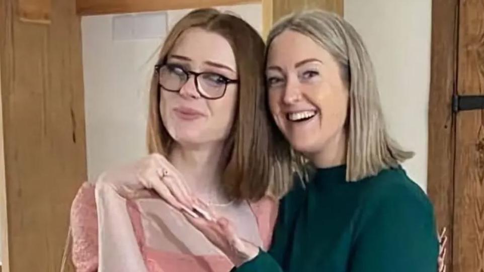 Brianna Ghey with long brown hair wearing black glasses and a peach dress with her mum Esther with bobbed blond hair wearing a dark green jumper. They are both smiling.