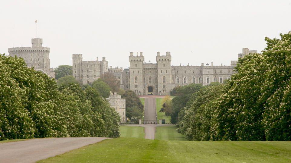 A long, tree-lined path leads to the grand entrance of Windsor Castle, with multiple towers and battlements. There's greenery on either side of the path and the sky above is overcast.