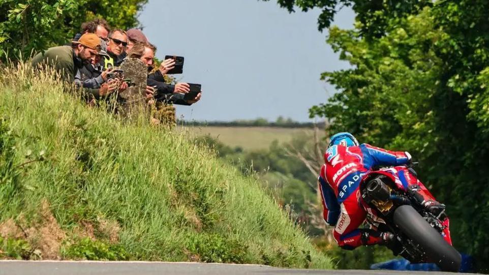 Dean Harrison in red and blue leathers riding away on the TT course. He is leaning over towards a grass embankment where a group of spectators are holding up mobile phones filming him.