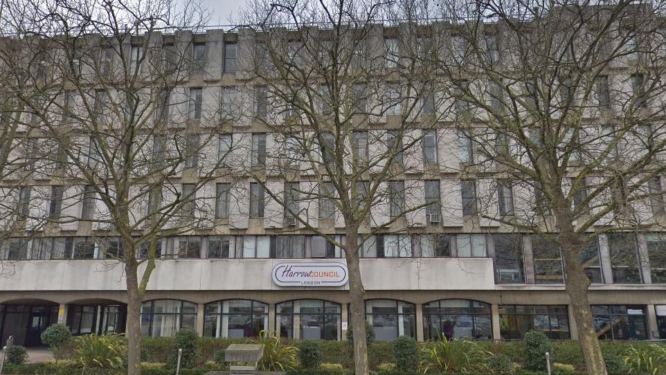 A tall concrete building with large windows, partly hidden behind bare trees. A sign above the entrance reads "Harrow Council, London.