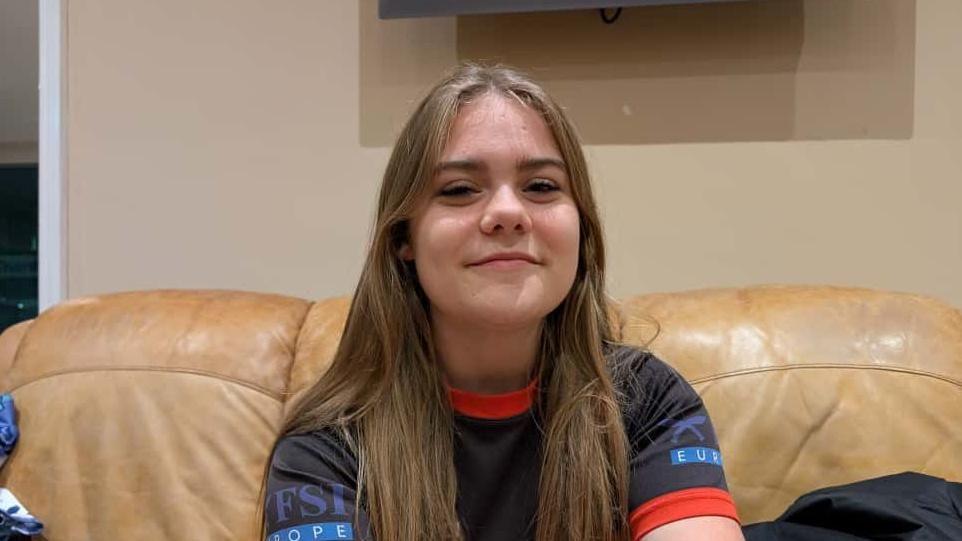 A girl from Chard Rugby Club with long fair hair wearing a navy kit with red trim. She is sitting on a beige leather sofa and she is smiling at the camera.
