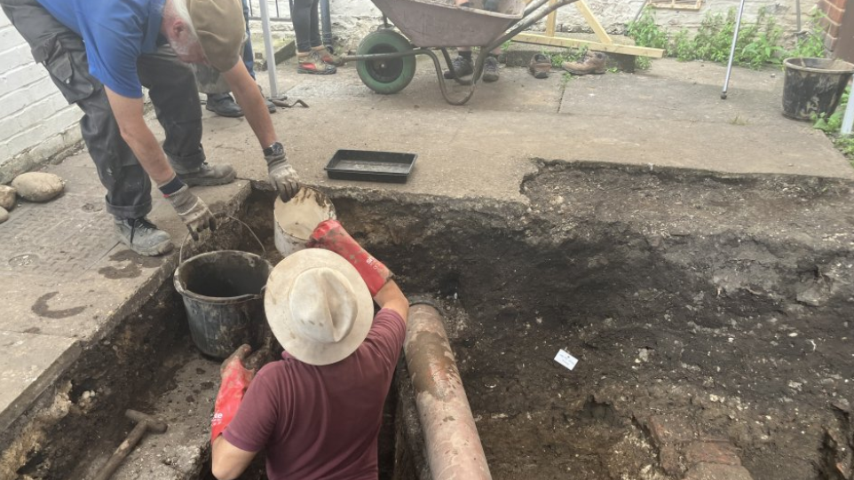 An archaeologist wearing a red polo shirt and a straw hat is standing in a trench. There is a man wearing a blue polo shirt and flat cap who is passing a white container and a black bucket to the person in the trench.  A wheelbarrow and a wooden handle (possibly a spade) are also visible. 