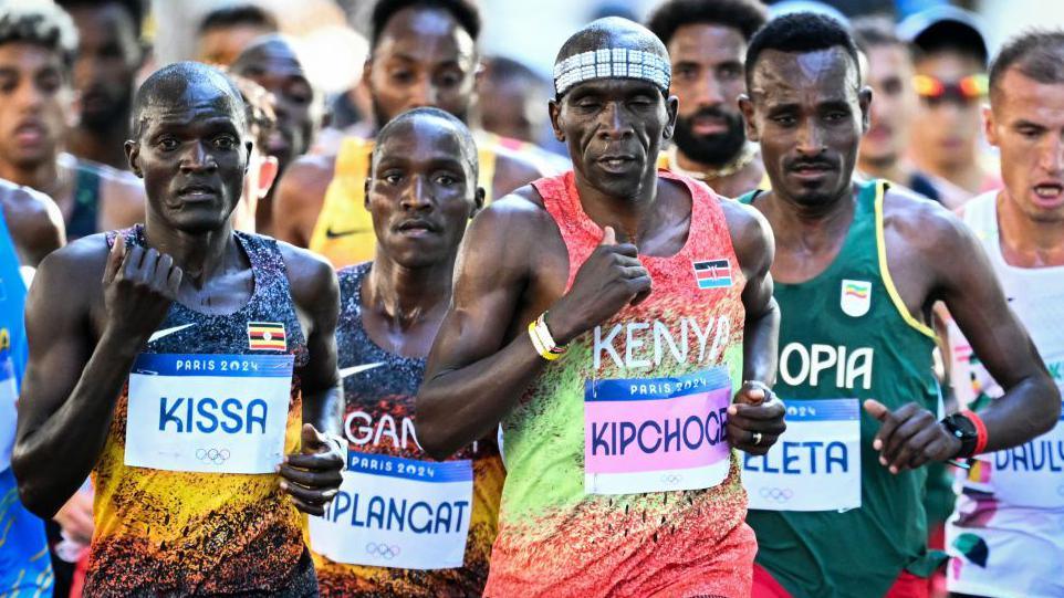 Eliud Kipchoge runs at the front of a pack of other athletes during the men's marathon at the Paris 2024 Olympic Games. He is second from the right in a group of four runners who dominate the image, wearing a vivid running Kenya running vest with dayglo orange and green colours, the word Kenya written in white and the flag on the left breast. He also has a pink bib with his name on it across his stomach, while he is also wearing a headband