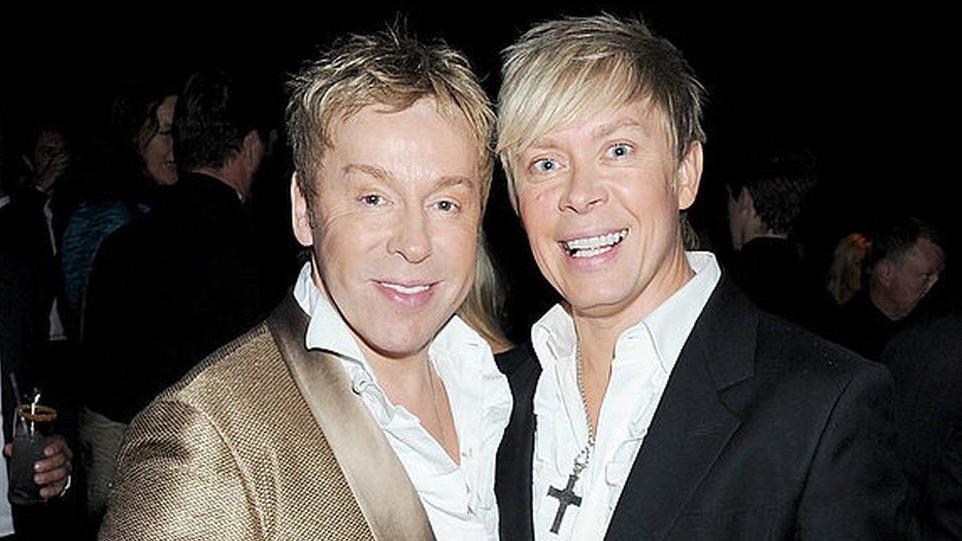 Royston Blythe, a man with short blonde hair and wearing a gold jacket and white shirt, standing next to Nick Malenko, a man with short blonde hair and wearing a black jacket and white shirt, with a crucifix necklace. Both are smiling at the camera.