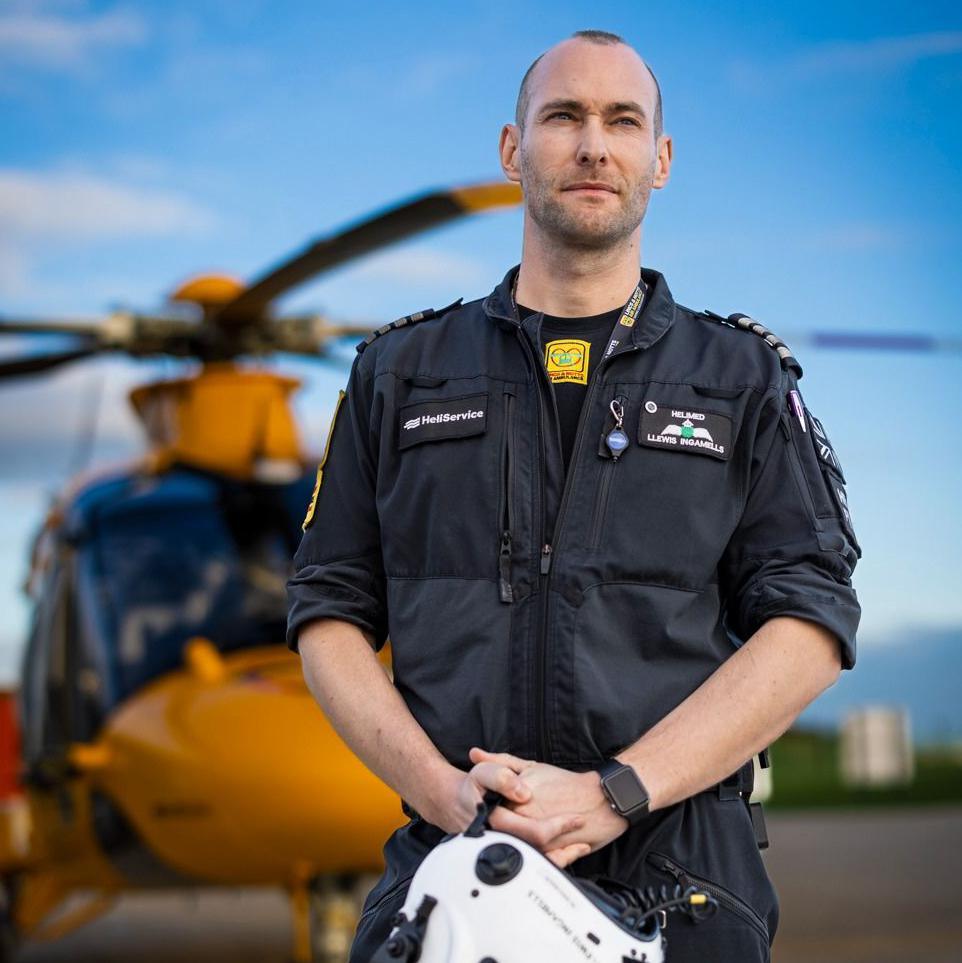 Llewis Ingamells wears a black flying suit and stands in front of the yellow air ambulance on a runway