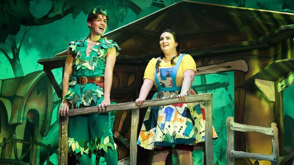 Bradley Riches on the left is dressed in a green tunic and cropped trousers with leaves attached, he is open mouthed as he is singing. 