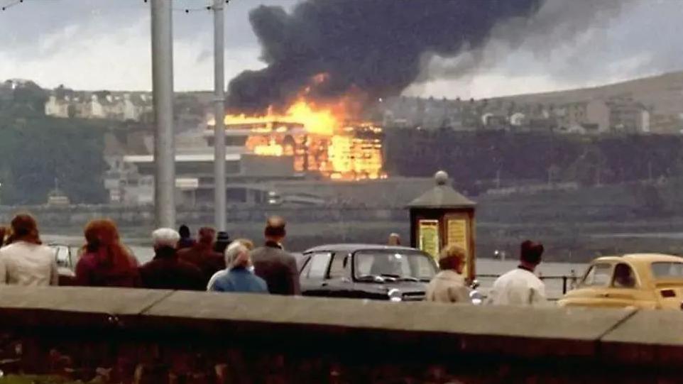 A crowd looks over the bay as a large complex that is ablaze, with billowing black smoke coming from the building. The cars and clothing are in keeping with the style of the 1970s.