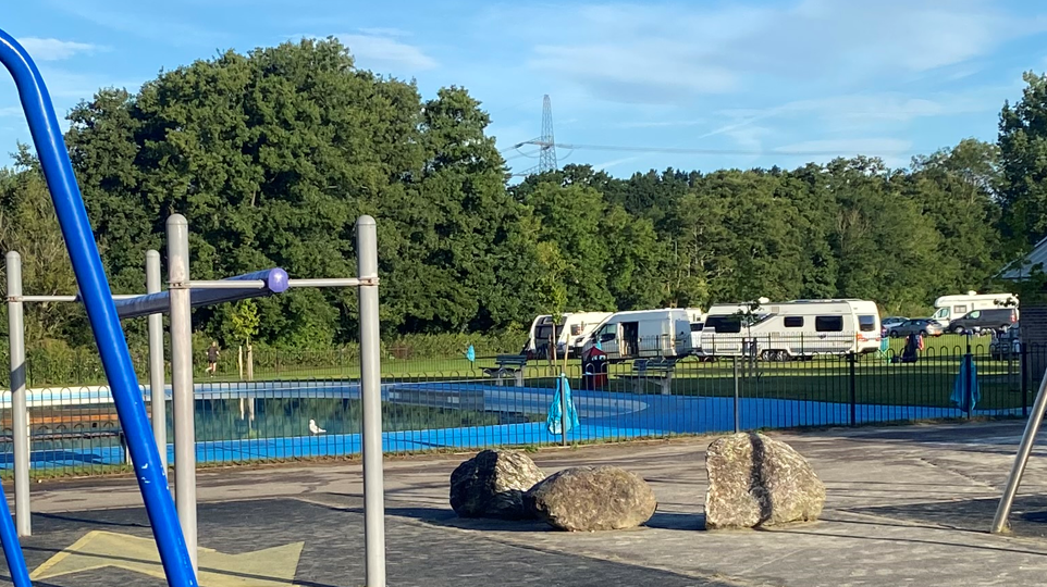 A large outdoor paddling pool in a children's park. There are several camper vans and cars on the grass to the side.