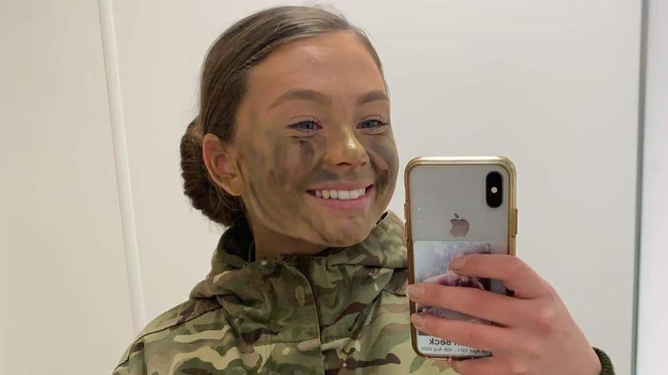 A mirror selfie of Gunner Beck wearing her camouflage military uniform, with her hair tied back into a slick low bun. She has camouflage green and brown face paint on and is smiling at the mirror.