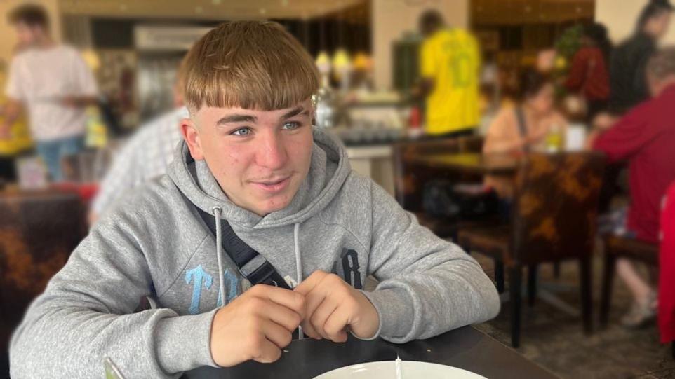 Kieron Hilling sitting in a restaurant, wearing a grey hoodie. He has short brown/blond hair and blue eyes. People are blurred in the background.