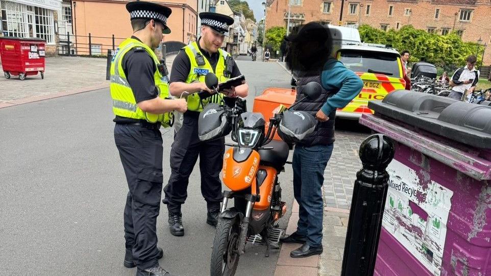 Two police officers speak to a Just Eat delivery driver who stands next to his electric cargo bike, which is black and orange with a large box on the back. The man's face is blurred out. He is wearing dark blue jeans, a dark grey gilet and a turquoise sweater. The officers are wearing chequered police hats, black combat trousers, black shirts and neon police vests. One holds an electronic tablet, which he is looking at. A police car is parked to one side and there are houses in the background.
